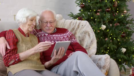 Glückliches-Seniorenpaar,-Das-Auf-Dem-Sofa-Sitzt-Und-Zur-Weihnachtszeit-Etwas-Interessantes-Auf-Dem-Digitalen-Tablett-Sieht