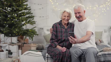 Glückliches-älteres-Paar,-Das-Auf-Dem-Bett-Sitzt-Und-Am-Weihnachtsmorgen-Einen-Videoanruf-Macht