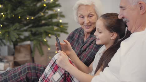 Glückliche-Großeltern-Mit-Ihrer-Kleinen-Enkelin-Sitzen-Auf-Dem-Sofa-Und-Nehmen-Am-Weihnachtsmorgen-Einen-Videoanruf-Entgegen