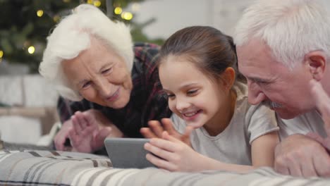 Glückliche-Großeltern-Mit-Ihrer-Kleinen-Enkelin,-Die-Auf-Dem-Bett-Liegen-Und-An-Weihnachten-Einen-Videoanruf-Tätigen