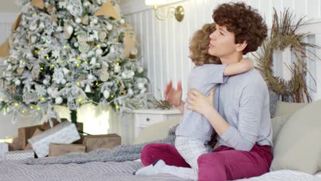 Loving-Mom-Hugging-Her-Little-Son-Sitting-On-Bed-In-A-Cozy-Bedroom-Decorated-With-A-Christmas-Tree