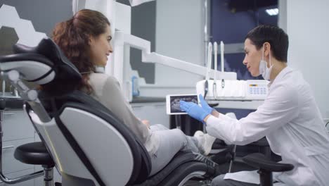 Mujer-Dentista-Con-Bata-Blanca-Y-Guantes-Sosteniendo-Una-Tableta-Y-Mostrando-Rayos-X-De-La-Mandíbula-A-Una-Paciente-Sentada-En-Una-Silla-Dental