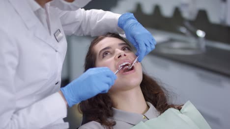 Zahnärztin-In-Weißem-Mantel,-Gesichtsmaske-Und-Handschuhen-Mit-Zahnsteinentferner-Und-Zahnspiegel-Bei-Der-Untersuchung-Der-Zähne-Junger-Patientinnen