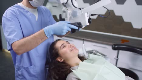 Male-Dentist-In-Scrubs,-Gloves-And-Face-Mask-Using-Dental-Microscope-And-Examining-Female-Patient's-Teeth
