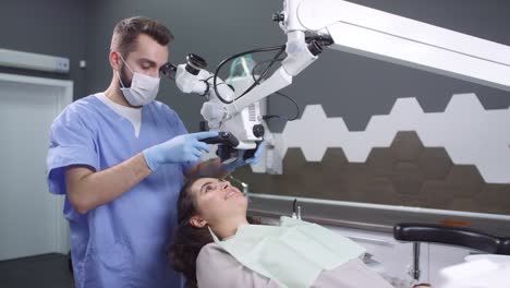 Male-Dentist-In-Scrubs,-Gloves-And-Face-Mask-Using-Dental-Microscope-And-Examining-Female-Patient's-Teeth-1