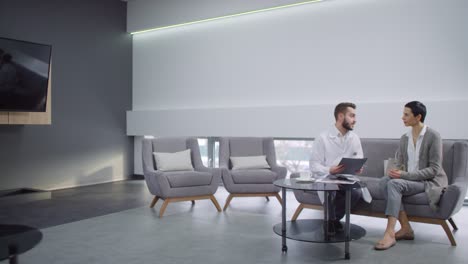 Male-Dentist-In-White-Coat-Sitting-On-Couch-In-Reception-Area-Of-Modern-Dental-Clinic-And-Discussing-Treatment-Options-With-Female-Patient