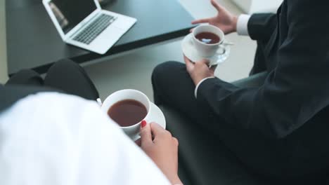Close-Up-Of-Two-Unrecognizable-Business-People-Holding-Coffee-Cup-And-Talking-While-They-Are-Sitting-On-Sofa-At-Office