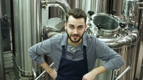 Handsome-Bearded-Brewery-Worker-In-Apron-Standing-At-Beer-Plant-And-Looking-At-Camera