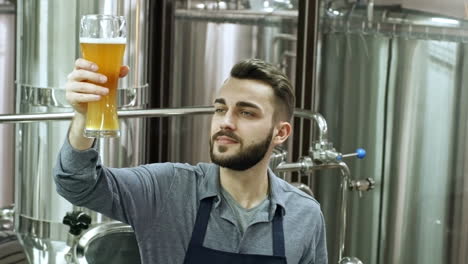 Young-Cheerful-Brewmaster-Looking-At-Glass-Of-Freshly-Brewed-Unfiltered-Beer-And-Smiling-At-Camera