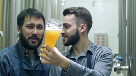 Zwei-Männliche-Mitarbeiter-Lächeln,-Betrachten-Frisch-Gebrautes-Unfiltriertes-Bier-Im-Glas-Und-Diskutieren-Es-In-Der-Brauerei