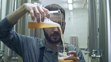 Young-Cheerful-Brewmaster-Looking-At-Glass-Of-Freshly-Brewed-Unfiltered-Beer-And-Pouring-It-Into-Another-Container