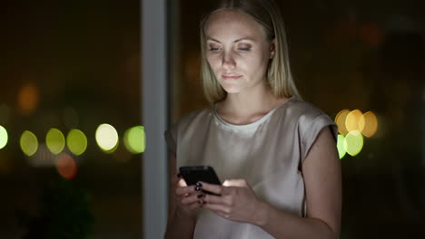 Nahaufnahme-Einer-Blonden-Frau,-Die-In-Der-Dunkelheit-Eine-Nachricht-Auf-Dem-Smartphone-Eingibt,-Mit-Defokussierten-Lichtern-Im-Hintergrund