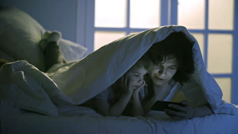 Cute-Little-Girl-And-Young-Mother-Lying-On-Bed-Under-Blanket-And-Reading-Fairy-Tale-With-Smartphone-Together
