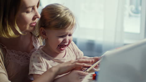 Young-Mother-Holding-Crying-Toddler-Girl-On-Her-Laps-And-Showing-Something-On-Mobile-Phone-While-Trying-To-Calm-Her-Down