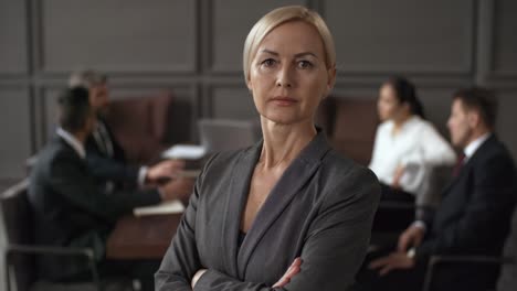Portrait-Of-A-Serious-Businesswoman-Standing-With-Crossed-Arms-And-Looking-At-Camera-While-In-Blurred-Background-Group-Of-Business-Executives-Sitting-Around-Table-During-Meeting