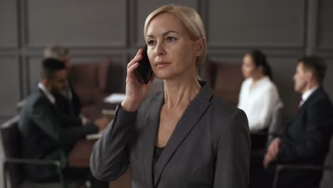 Portrait-Of-A-Serious-Businesswoman-Standing-And-Speaking-On-Mobile-Phone,-While-In-Blurred-Background-Group-Of-Business-Executives-Sitting-Around-Table-During-Meeting