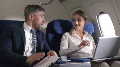 Cheerful-Businesswoman-With-Laptop-And-Bearded-Businessman-Holding-Clipboard-With-Documents-Discussing-Work-And-Shaking-Hands-While-Traveling-By-Plane