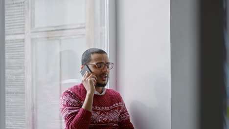 Arabischer-Mann-Mit-Brille-Und-Weihnachtspullover-Sitzt-Im-Studio-Und-Telefoniert-Mit-Einem-Fenster-Im-Hintergrund