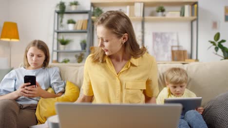 Female-Teacher-Having-Online-Class-At-Home-And-Showing-Charts-And-Statistics-While-Her-Children-Sitting-On-Sofa-Behind-Her-Using-Electronic-Devices