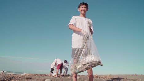 Smiling-Young-Indonesian-Boy-Picking-Up-Flip-Flop-And-Putting-It-In-Special-Trash-Bag-While-His-Team-Collecting-Garbage-In-Background-Too