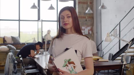 Portrait-Of-Beautiful-Young-Fashion-Designer-Holding-Her-Sketchbook-And-Smiling-At-Camera-In-Sewing-Workshop-While-Her-Colleagues-Working-In-The-Background