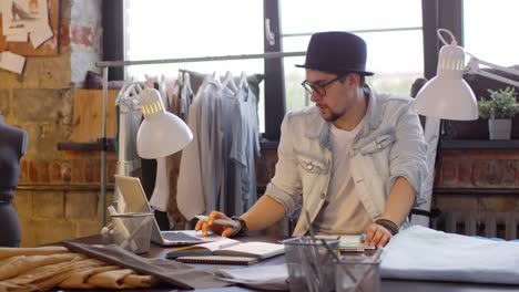 Young-Male-Fashion-Designer-Sitting-At-Desk,-Using-Laptop-And-Drawing-Sketch-Of-New-Dress-In-Modern-Sewing-Studio-1