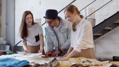 Team-Of-Male-And-Two-Female-Designers-Discussing-Fashion-Sketch-At-Worktable-In-Sewing-Studio