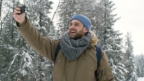Hombre-Guapo-Alegre-Videollamada-En-El-Teléfono-Inteligente,-Saludando-A-La-Cámara,-Sonriendo-Y-Contando-Sobre-La-Hermosa-Naturaleza-Invernal-En-El-Parque-Nacional-Mientras-Camina-En-Un-Día-Nevado