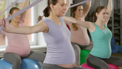 Vista-Lateral-De-Cuatro-Mujeres-Embarazadas-En-Forma-Sentadas-En-Pelotas-De-Fitness-De-Estabilidad-Y-Usando-Bandas-Para-Ejercicios-De-Calentamiento