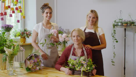 Middle-Aged-Flower-Shop-Proprietor-Sitting-At-Wooden-Table-With-Exquisite-Floral-Display-And-Posing-With-Two-Young-Florists,-With-Bunches-Of-Flowers-And-Bouquets-In-Background