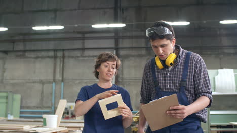 Man-And-Woman-Standing-Together-In-Carpentry-Shop-And-Discussing-New-Project-While-Their-Colleague-Carrying-Wooden-Plank-On-Background
