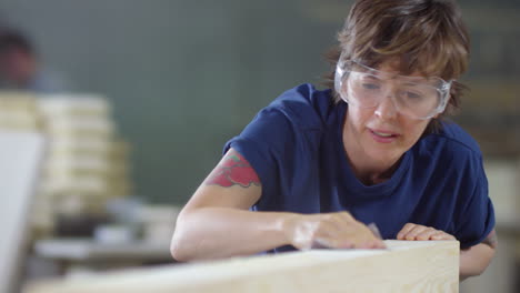 Woman-In-Safety-Goggles-Sanding-Timber-With-Sandpaper-And-Then-Blowing-Dust-Away