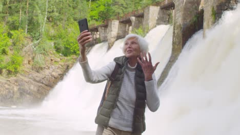 Excited-Elderly-Woman-With-Grey-Hair-Standing-In-Front-Of-Dam-Spillway-And-Video-Calling-Someone-On-Mobile-Phone