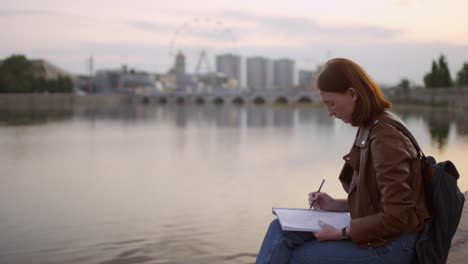 Pretty-Ginger-Young-Woman-With-Freckles-Sitting-On-Embankment-And-Drawing-Cityscape