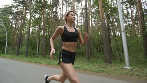 Side-View-Of-Young-Woman-In-Sports-Top-And-Shorts-Running-Along-Road-In-Park-In-Morning