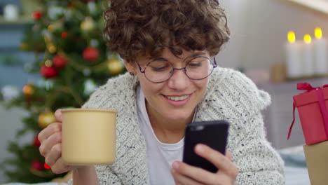 Woman-With-Short-Curly-Hair-Lying-On-The-Floor-At-Home-Using-Smartphone-In-A-Room-With-Christmas-Decorations