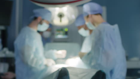 Blurred-View-Of-Four-Medical-Professionals-In-Sanitary-Clothes-Performing-Surgery-On-Patient-At-Hospital