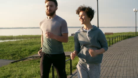 Sporty-Young-Man-And-Woman-Smiling-And-Talking-While-Running-Together-On-Riverside-Promenade-In-The-Morning