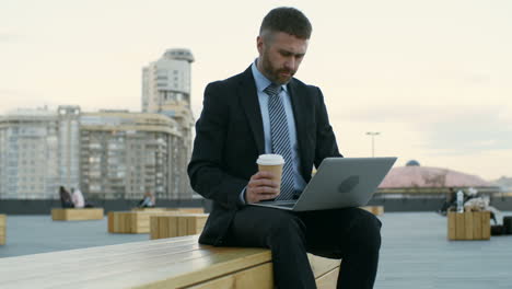 Geschäftsmann-Sitzt-Draußen-Auf-Einer-Holzbank-Mit-Laptop-Auf-Den-Knien,-Tippt-Auf-Der-Tastatur-Und-Trinkt-Kaffee