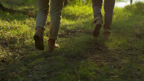 Rear-View-Following-Shot-Of-Legs-Of-Male-And-Female-Hikers-Walking-On-Green-Grass-Through-The-Forest