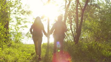 Loving-Tourist-Couple-Holding-Hands,-Smiling-And-Talking-While-Walking-Together-Through-Green-Forest-At-Sunset