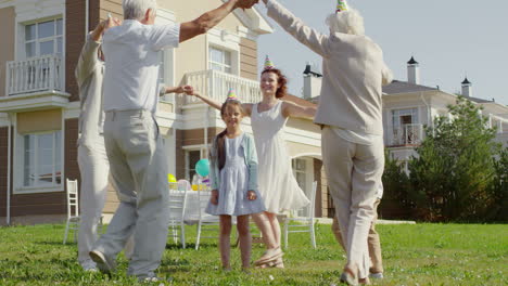 Feliz-Familia-Multigeneracional-Con-Sombreros-De-Fiesta-Tomándose-De-La-Mano-Y-Bailando-Alrededor-De-Una-Niñita-Mientras-Celebraba-Al-Aire-Libre-En-El-Patio-Trasero-El-Día-Soleado