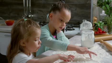 Two-Little-Sisters-Of-Primary-School-Age-Cooking-In-Kitchen-And-Drawing-With-Fingers-On-Floured-Surface