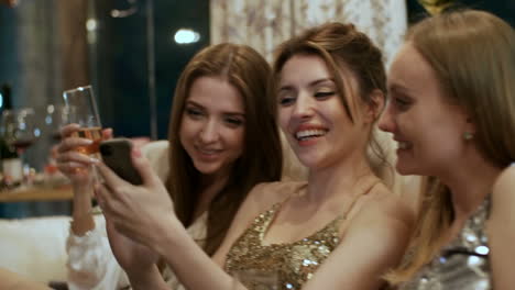 Three-Relaxed-Girlfriends-Sitting-Together-At-Party,-Checking-Updates-On-Smartphone-And-Laughing-Animatedly