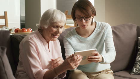 Caring-Female-Volunteer-Showing-Retired-Woman-How-To-Use-Tablet-Computer-Application
