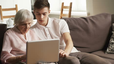 ältere-Frau-Und-Junger-Mann-Sitzen-Auf-Dem-Sofa-Im-Wohnzimmer-Und-Unterhalten-Sich,-Während-Sie-Einen-Laptop-Verwenden
