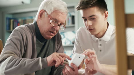 Junge-Pflegekraft,-Die-Mit-Einem-älteren-Mann-Mit-Brille-Spricht-Und-Ihm-Dabei-Hilft,-Die-Smartphone-Anwendung-Zu-Verwenden