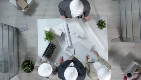 Above-View-Of-Two-Architects-In-Safety-Helmets-Standing-At-Table-Shaking-Hands-And-Having-Talk-Concerning-Blueprint