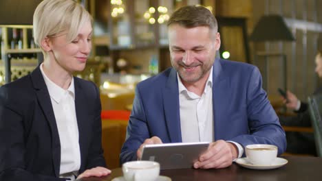 Middle-Aged-Businessman-Showing-Something-On-Tablet-To-His-Female-Colleague-While-They-Sitting-In-Bar-At-Table