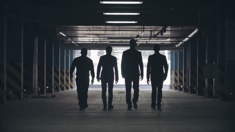 Slow-Motion-Locked-Down-Rear-View-Of-Silhouettes-Of-Four-Men-In-Black-Suits-Confidently-Walking-Away-From-Camera-Towards-Exit-Of-Parking-Lot